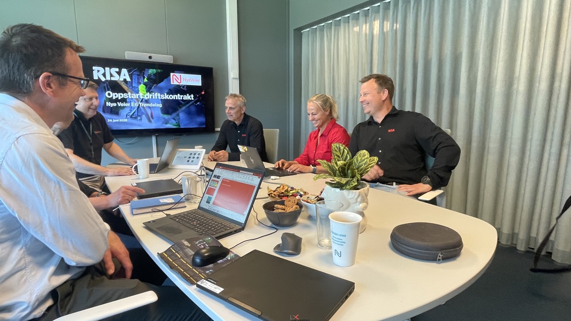 Personer som jobber sammen på møterom. Presentasjon på skjerm. Åpne laptoper. Smil og latter.